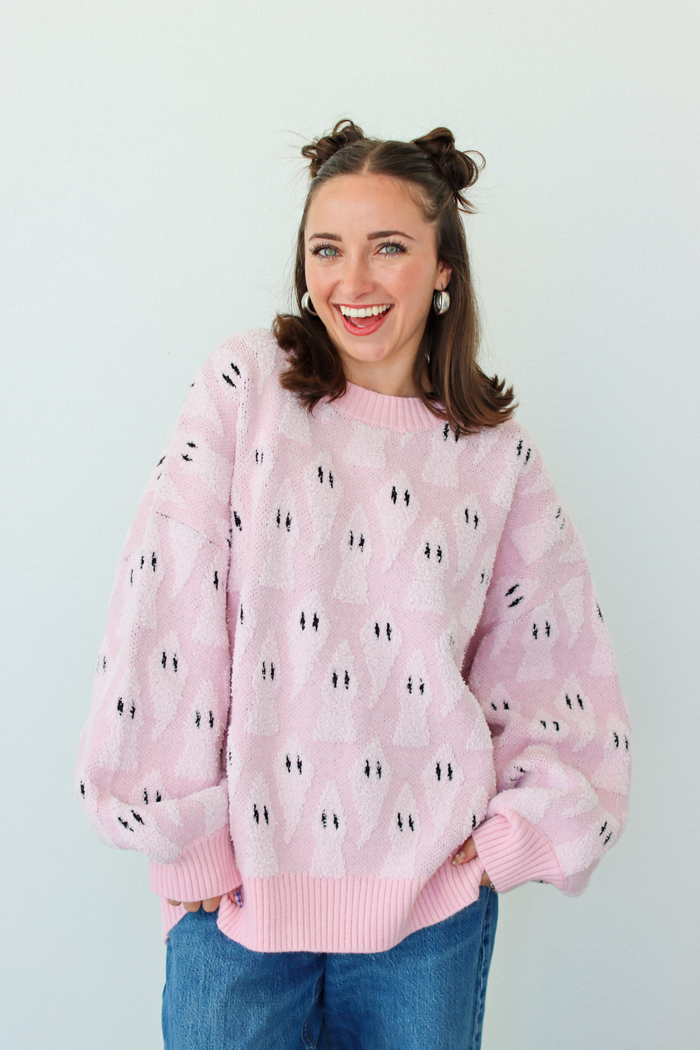 A smiling woman with her hair in two buns wears a light pink fuzzy knit sweater with a repeating black and white ghost pattern, paired with blue jeans, against a plain light gray background.