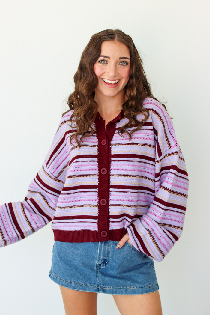 A woman modeling a pink and dark purple striped knit cardigan with a button-front closure, paired with a denim mini skirt, against a plain white background.