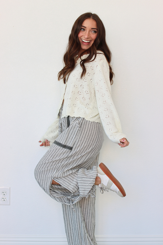 A woman modeling a cream eyelet knit cardigan over a white top and gray-and-white vertically striped wide-leg pants, standing against a plain white wall.